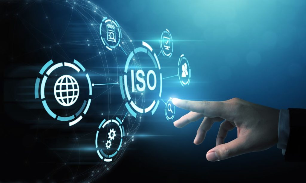 una mano humana tocando una interfaz digital con iconos tecnológicos flotantes. En el centro se destaca el texto “ISO”, representando los estándares internacionales de calidad y gestión. A su alrededor aparecen símbolos relacionados con la conectividad global, la configuración, el calendario y la gestión de usuarios, todo sobre un fondo azul futurista que evoca innovación y certificación tecnológica.