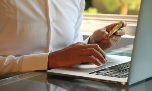 Fotografía en primer plano de las manos de una persona que está sosteniendo una tarjeta de crédito frente a su ordenador portátil, a punto de realizar una compra online.