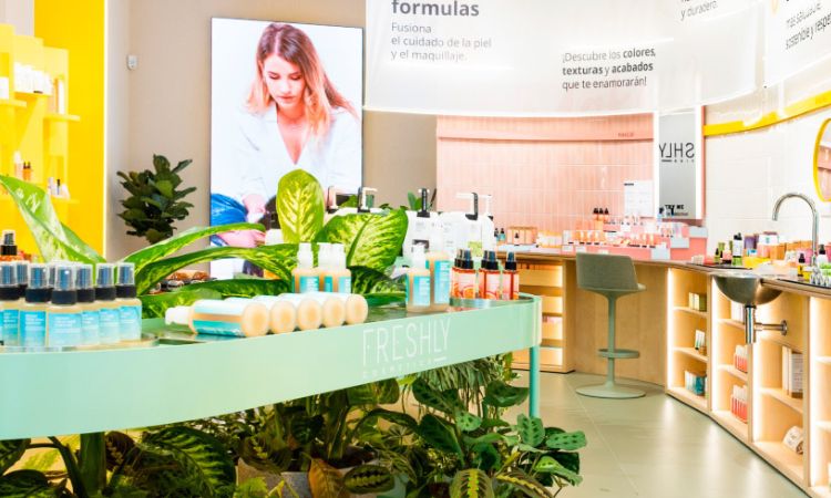 Exhibición de productos cosméticos, organizados en una tienda moderna y colorida con plantas decorativas; el ambiente es acogedor y profesional. Texto visible: "fórmulas", "pruebas en cabina, resultados visibles en tu piel".