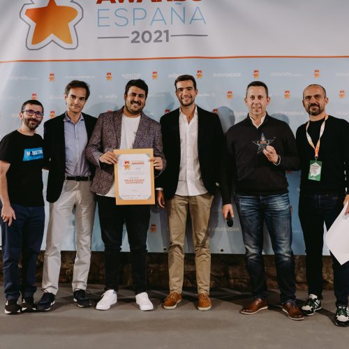 Grupo de seis hombres posando con un certificado en un evento de premios. Fondo: pancarta con estrella y texto "PREMIOS ESPAÑA 2021". Evento empresarial.