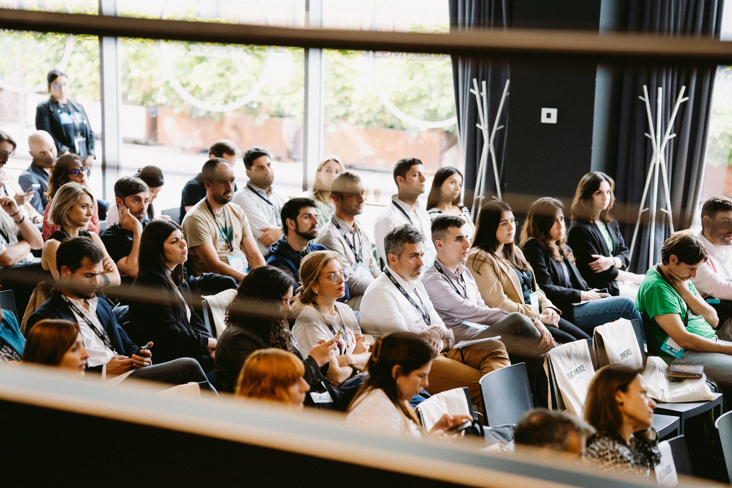 Personas asistiendo a conferencia en un auditorio. Todos están atentos, algunos toman notas. Ambiente iluminado por grandes ventanales. Marketing, networking.