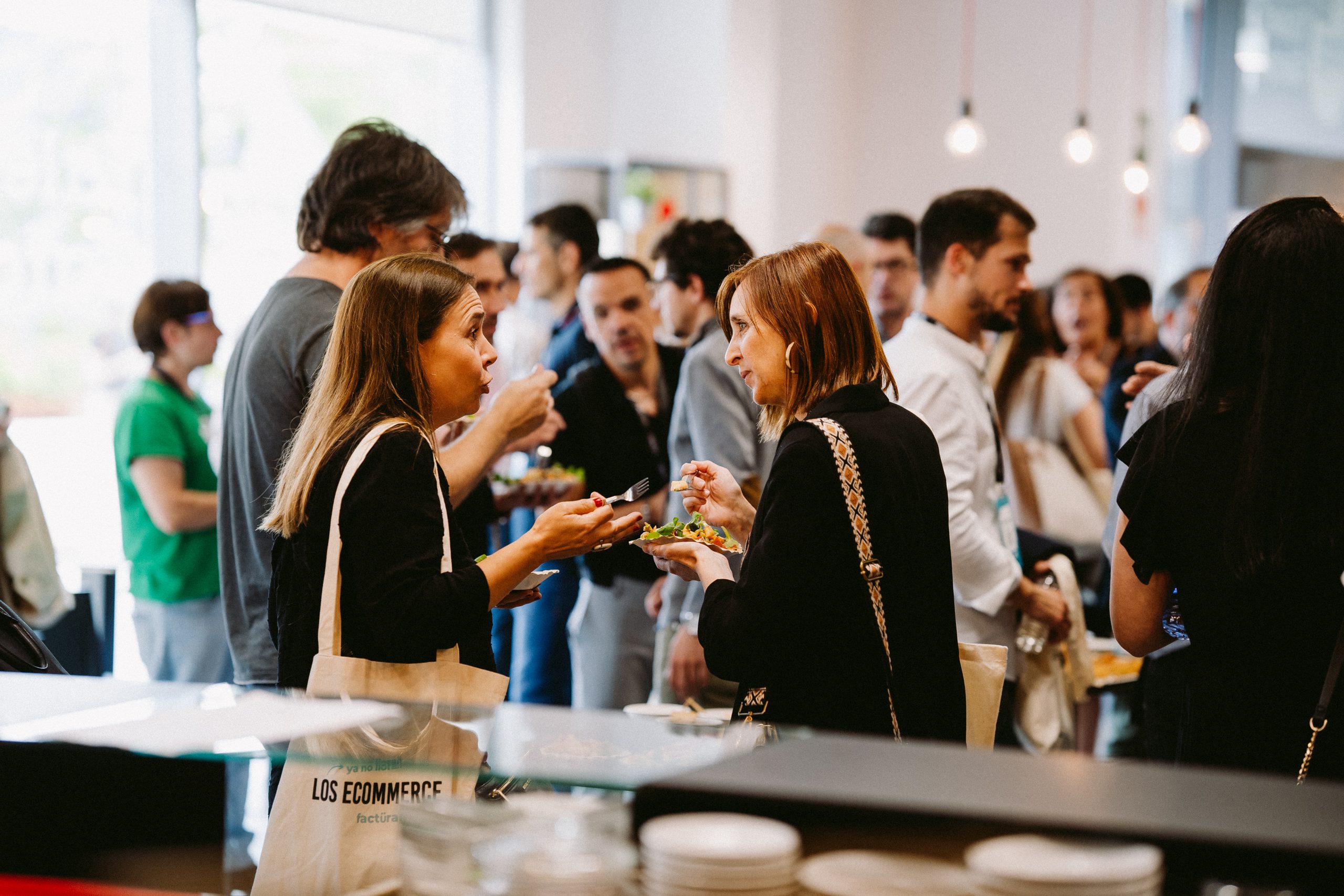 Personas conversan animadamente mientras comen en un evento de networking. El texto en una bolsa dice: “LOS ECOMMERCE”. Ambiente social y profesional.
