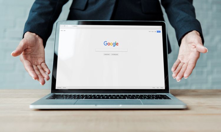 Un ordenador portátil colocado sobre una mesa muestra el buscador de Google en su pantalla. Tras el ordenador hay una persona que estira sus brazos hacia delante, con las palmas hacia el frente, enmarcando y resaltando el ordenador