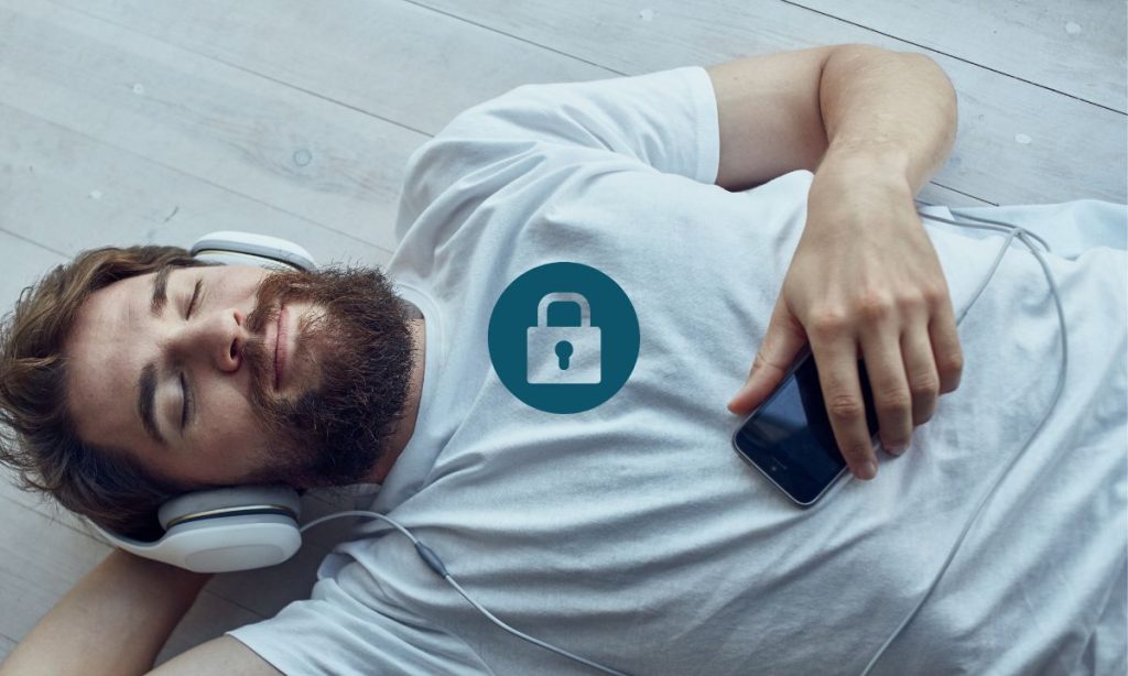 Imagen que muestra a un hombre con barba acostado sobre el suelo, usando audífonos y sosteniendo un teléfono móvil en su mano derecha. Está relajado. Sobre él aparece superpuesto un icono gráfico de un candado cerrado, lo que simboliza visualmente la temática de privacidad, protección de datos o seguridad digital.